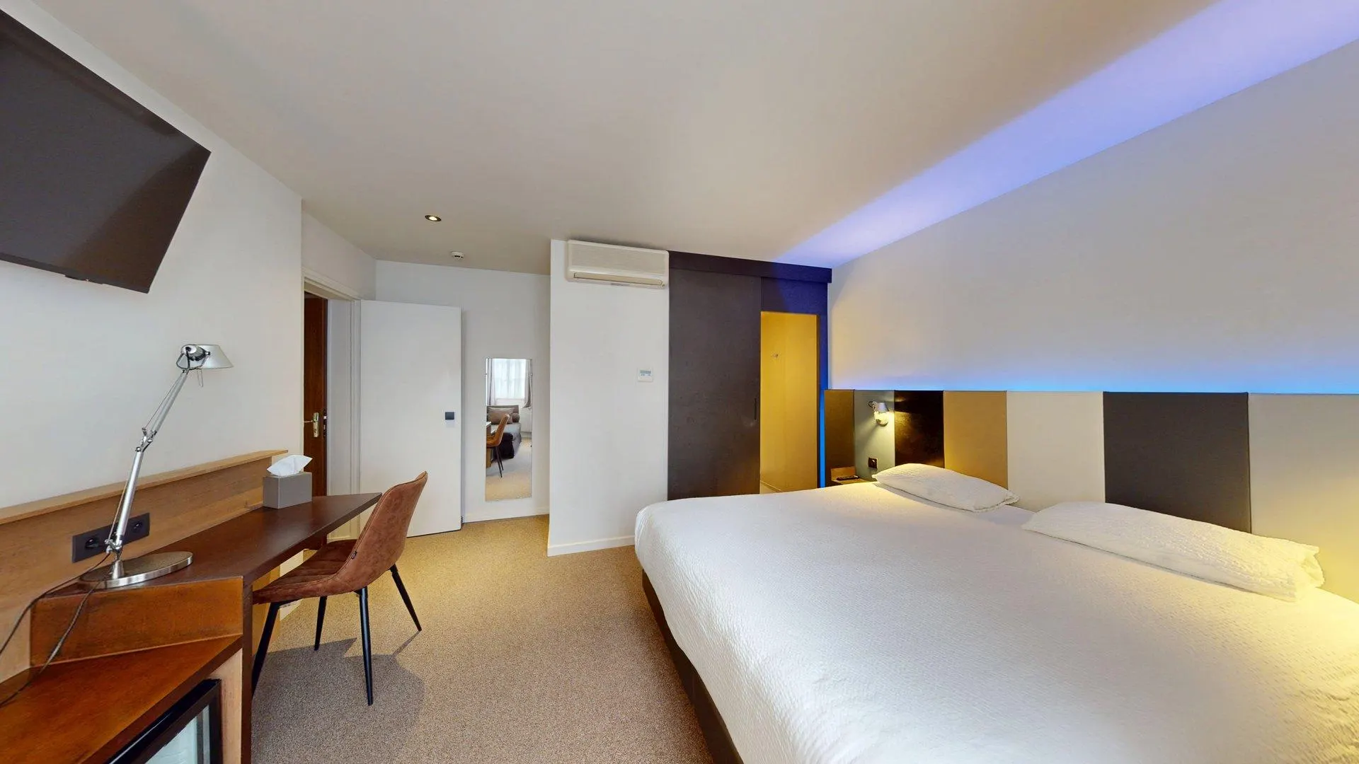 Chambre moderne à l'Hôtel de Harlay avec un grand lit, un bureau et une chaise en bois, une télévision murale, un miroir sur toute la longueur et un éclairage bleu ambiant au-dessus du lit - une expérience d'hébergement parfaite à Compiègne.