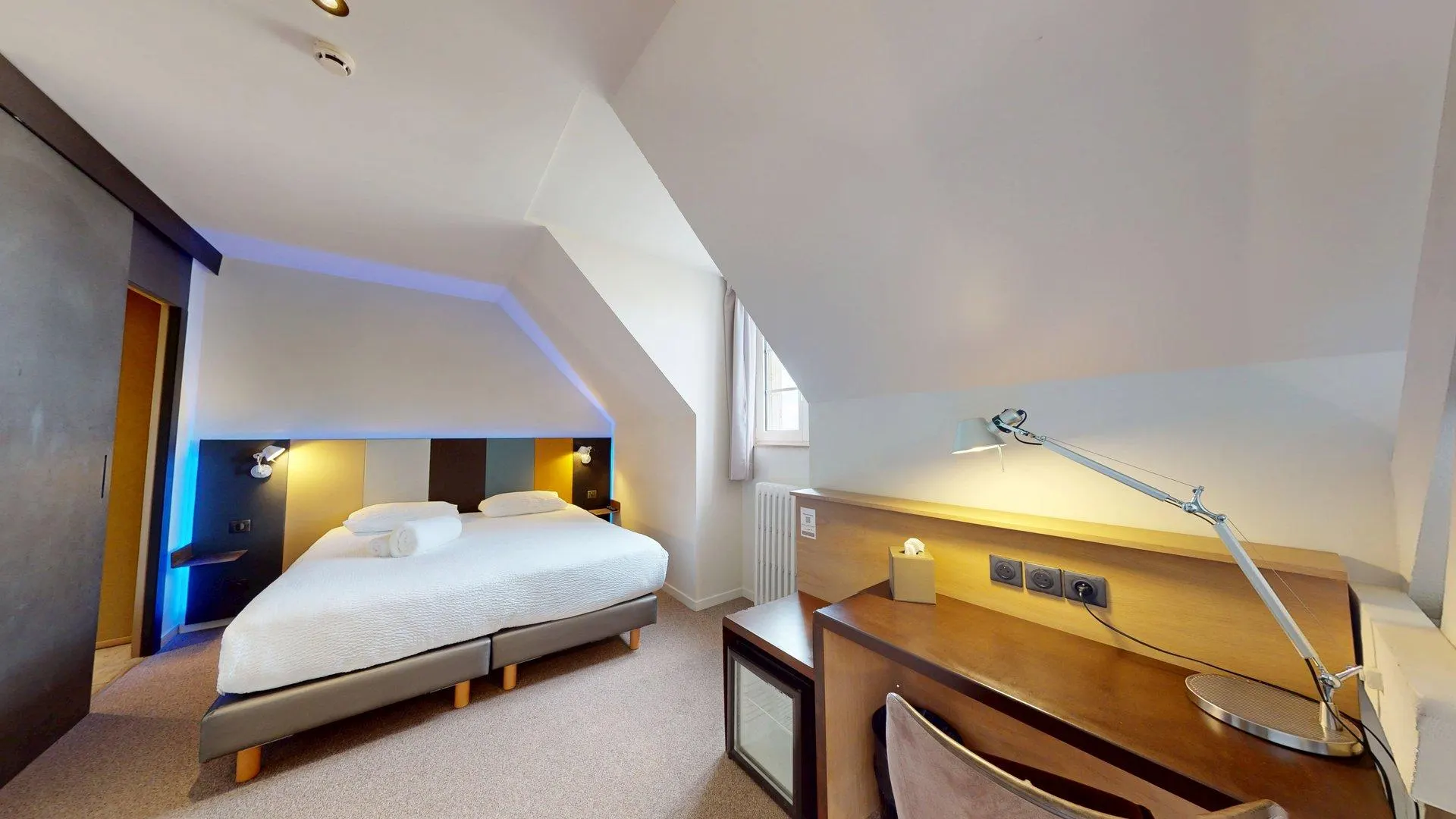 Chambre confortable à l'Hôtel de Harlay à Compiègne, comprenant un lit double avec des serviettes fraîches, un bureau et une lampe, un mini-réfrigérateur, et un plafond incliné avec une fenêtre - parfait pour votre expérience de l'Hébergement Compiègne.