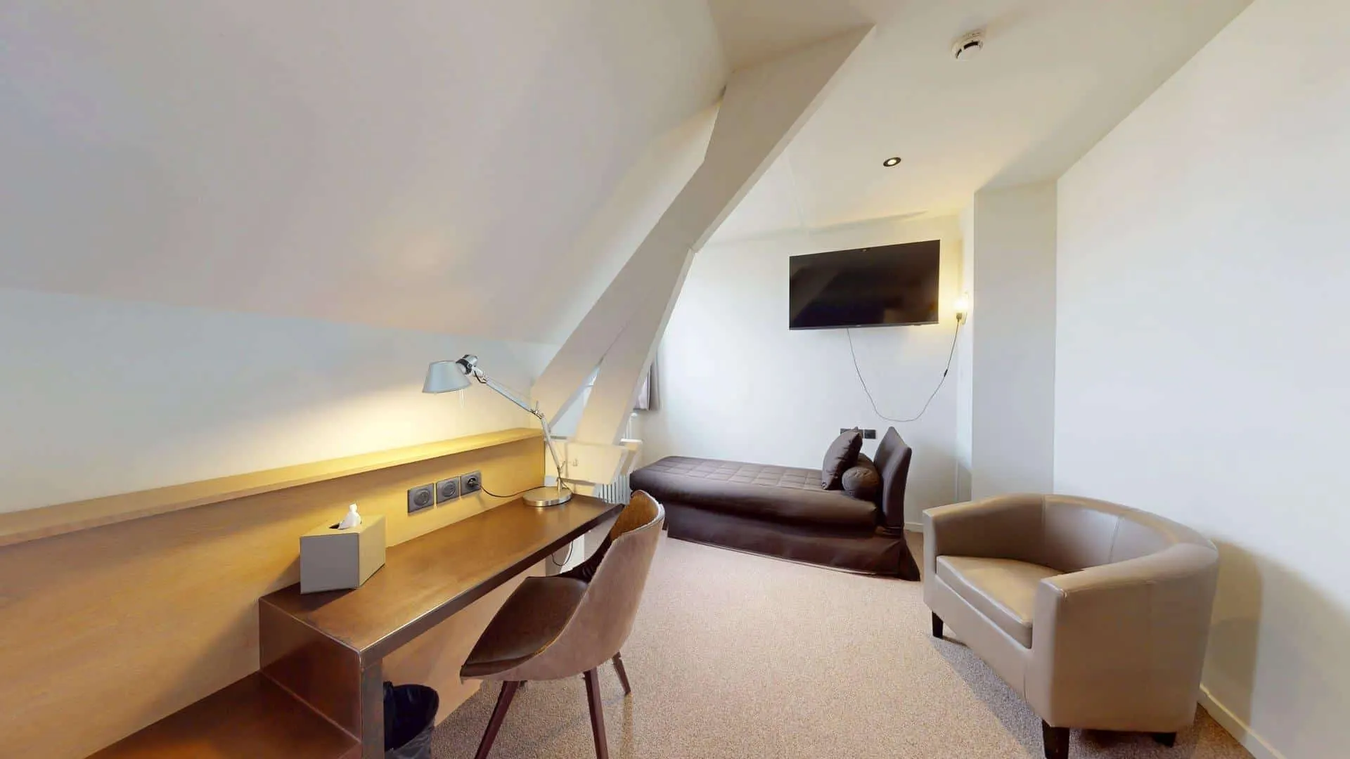 Une chambre d'hôtel moderne à l'Hôtel de Harlay avec un plafond incliné, comprenant un bureau avec une chaise et une lampe, un fauteuil, une chaise longue et une télévision murale - idéal pour un hébergement confortable à Compiègne.