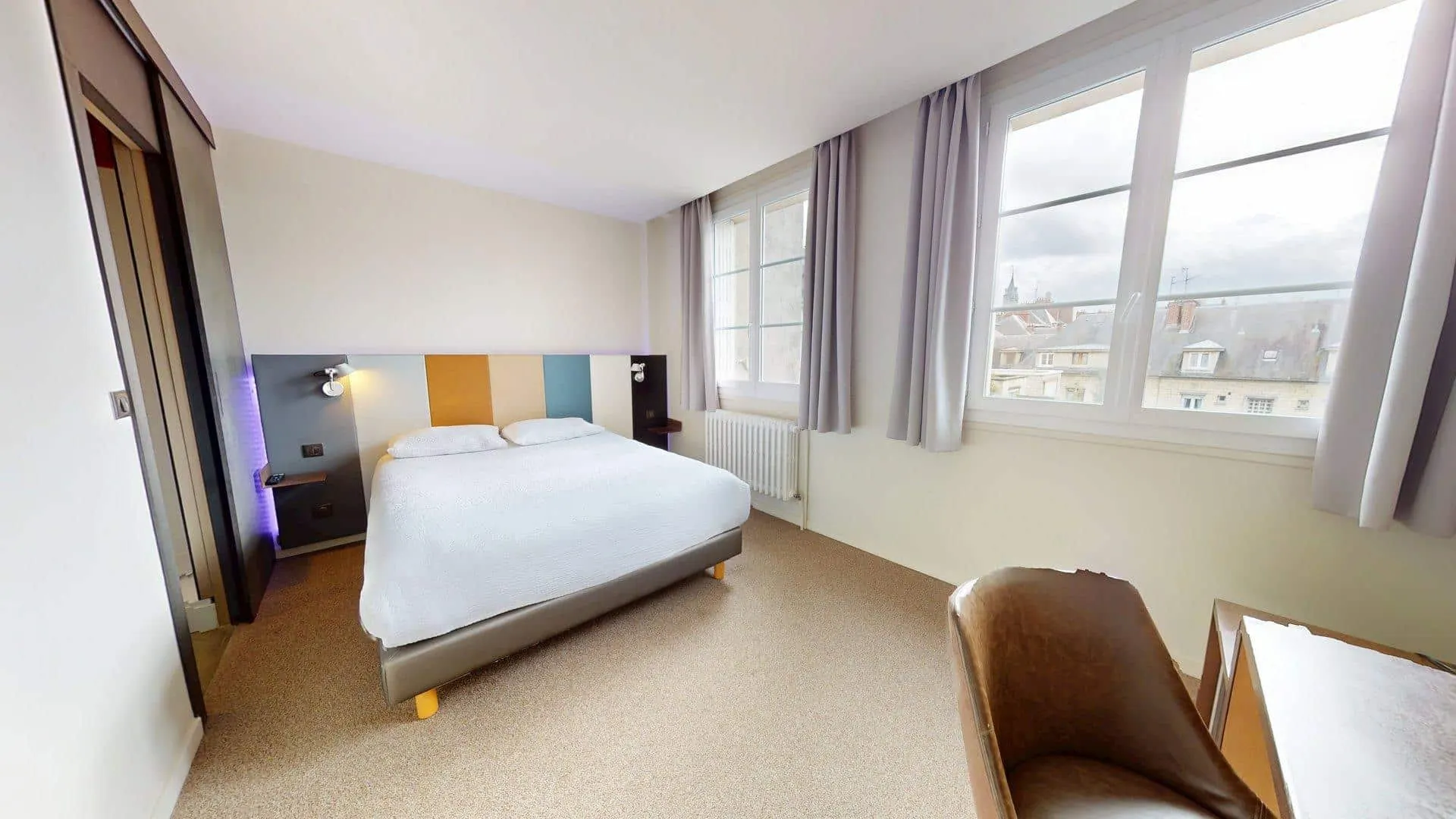 Une petite chambre de l'Hôtel de Harlay avec un lit double, une tête de lit multicolore, un bureau, une chaise brune, de grandes fenêtres avec des rideaux, et une vue sur les toits voisins - parfait pour un hébergement confortable à Compiègne.