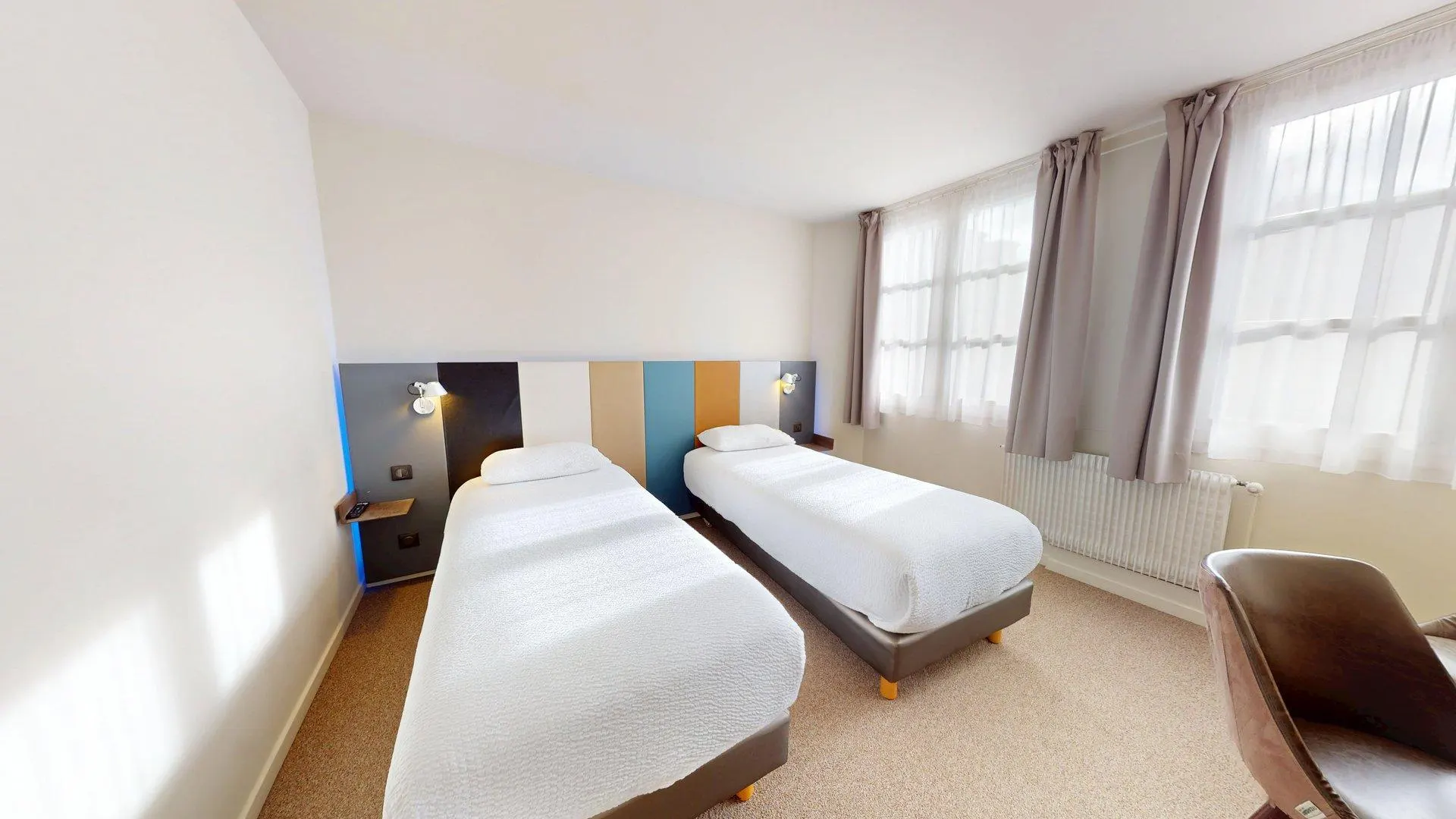 Une chambre d'hôtel à l'Hôtel de Harlay avec deux lits simples, une tête de lit colorée, des liseuses murales, deux fenêtres avec des rideaux transparents et une chaise marron - un choix parfait pour Hébergement Compiègne.