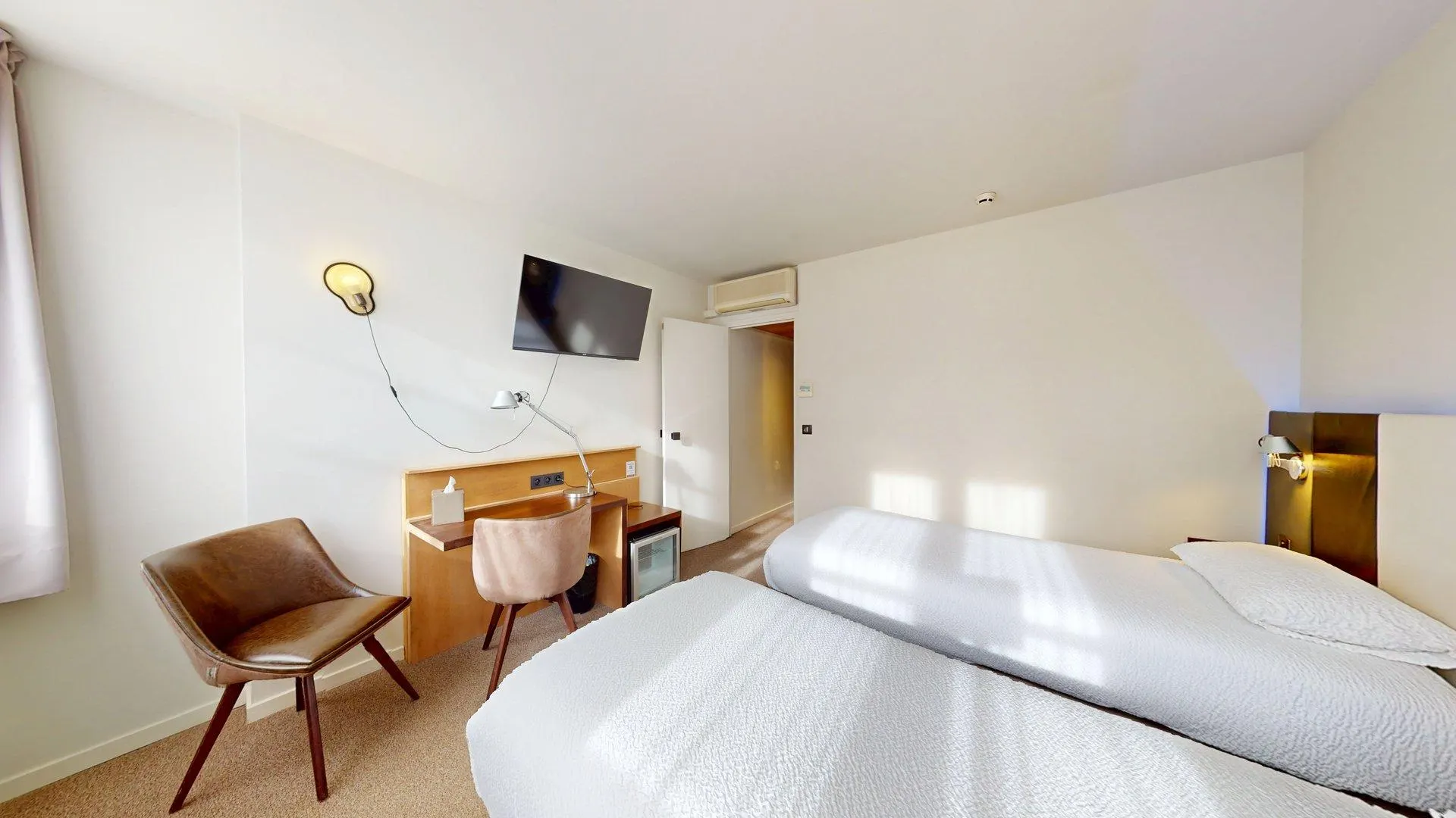 Chambre d'hôtel à l'Hôtel de Harlay avec deux lits simples, un bureau et une chaise, une télévision murale, un petit réfrigérateur et un décor neutre. La lumière du soleil pénètre par une fenêtre sur la gauche - idéal pour un hébergement confortable à Compiègne.