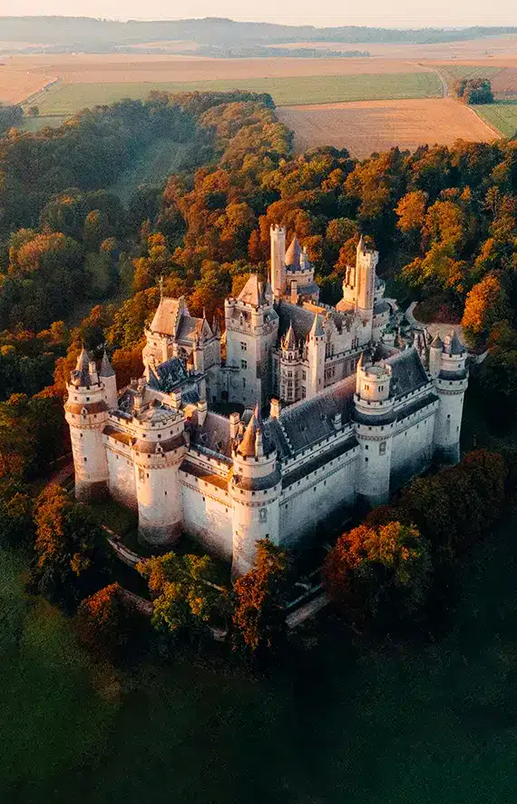 Vue aérienne d'un grand château en pierre, rappelant l'Hôtel de Harlay, entouré d'arbres denses et de champs, éclairé par la lumière chaude du soleil.