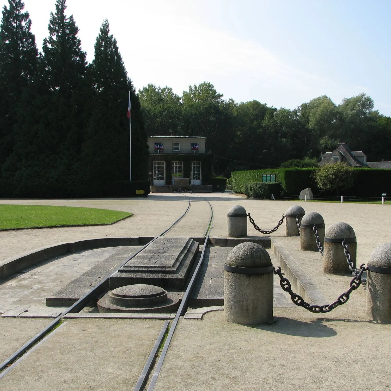 Monument en pierre avec des chaînes et des voies ferrées encastrées menant à un petit bâtiment, entouré d'arbres et de terrains ouverts sous un ciel clair - rappelant le charme historique trouvé près de l'Hôtel à Compiègne.