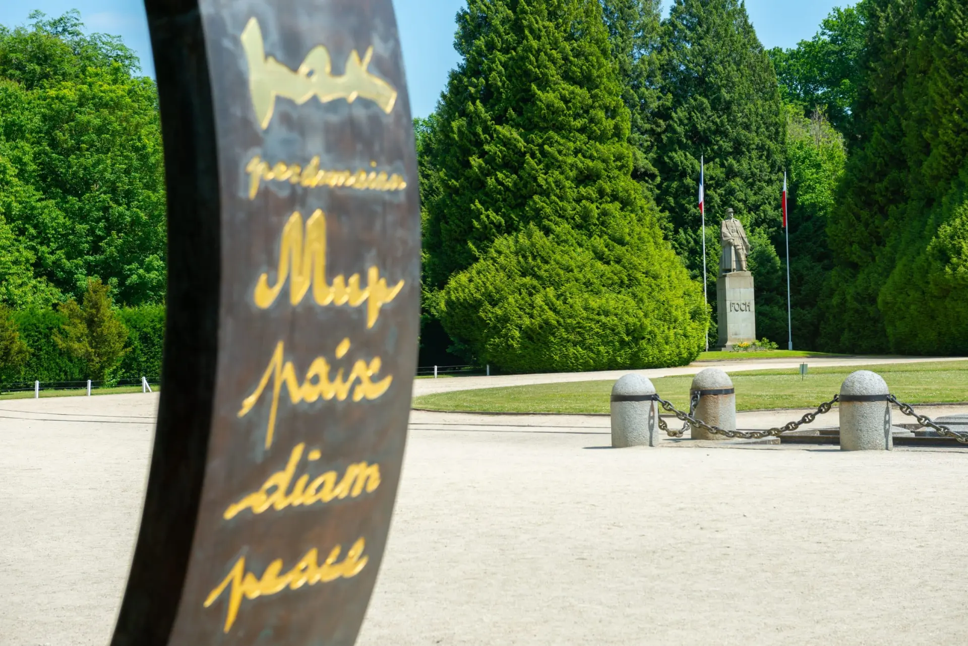 Gros plan sur une sculpture portant le mot "paix" en plusieurs langues, avec une statue et de grands arbres en arrière-plan, près de l'Hôtel de Harlay.