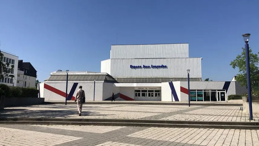 Une personne se dirige vers un bâtiment moderne et blanc, l'Espace Jean Legendre, sous un ciel bleu clair, avec des accents géométriques colorés sur la façade, à quelques pas des meilleures options d'Hébergement Compiègne, comme l'Hôtel de Harlay.