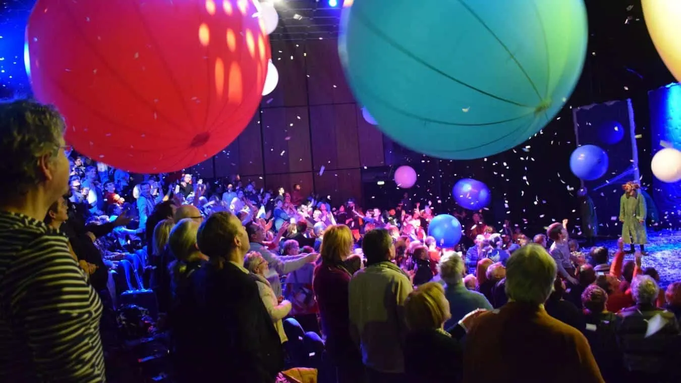 Une foule assise dans un théâtre près de l'Hôtel à Compiègne interagit avec de gros ballons colorés et des confettis qui tombent pendant une représentation animée sur scène.