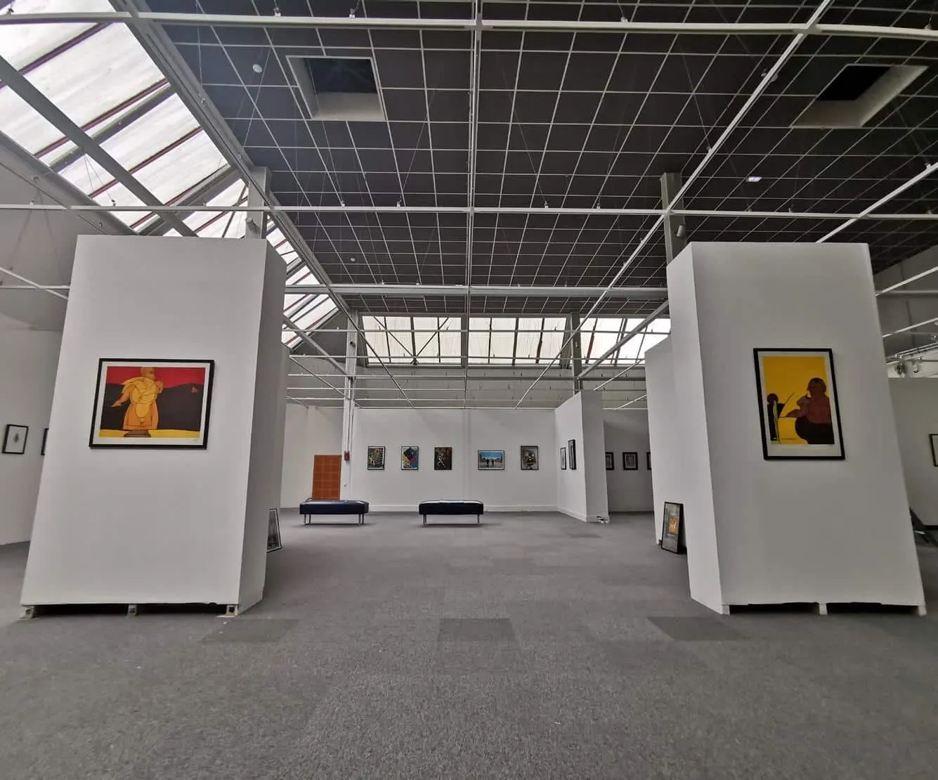 Intérieur spacieux d'une galerie d'art avec des murs blancs, des œuvres d'art encadrées, une moquette grise, des bancs et un plafond quadrillé avec des lucarnes - rappelant l'ambiance élégante de l'Hôtel de Harlay à Compiègne.