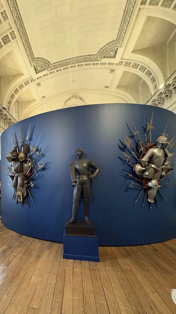 La statue d'un homme en uniforme se tient devant un présentoir circulaire bleu orné d'armures et d'armes, à l'intérieur d'un musée aux plafonds ornés et aux planchers en bois, à quelques pas de l'Hébergement Compiègne comme de l'Hôtel de Harlay.