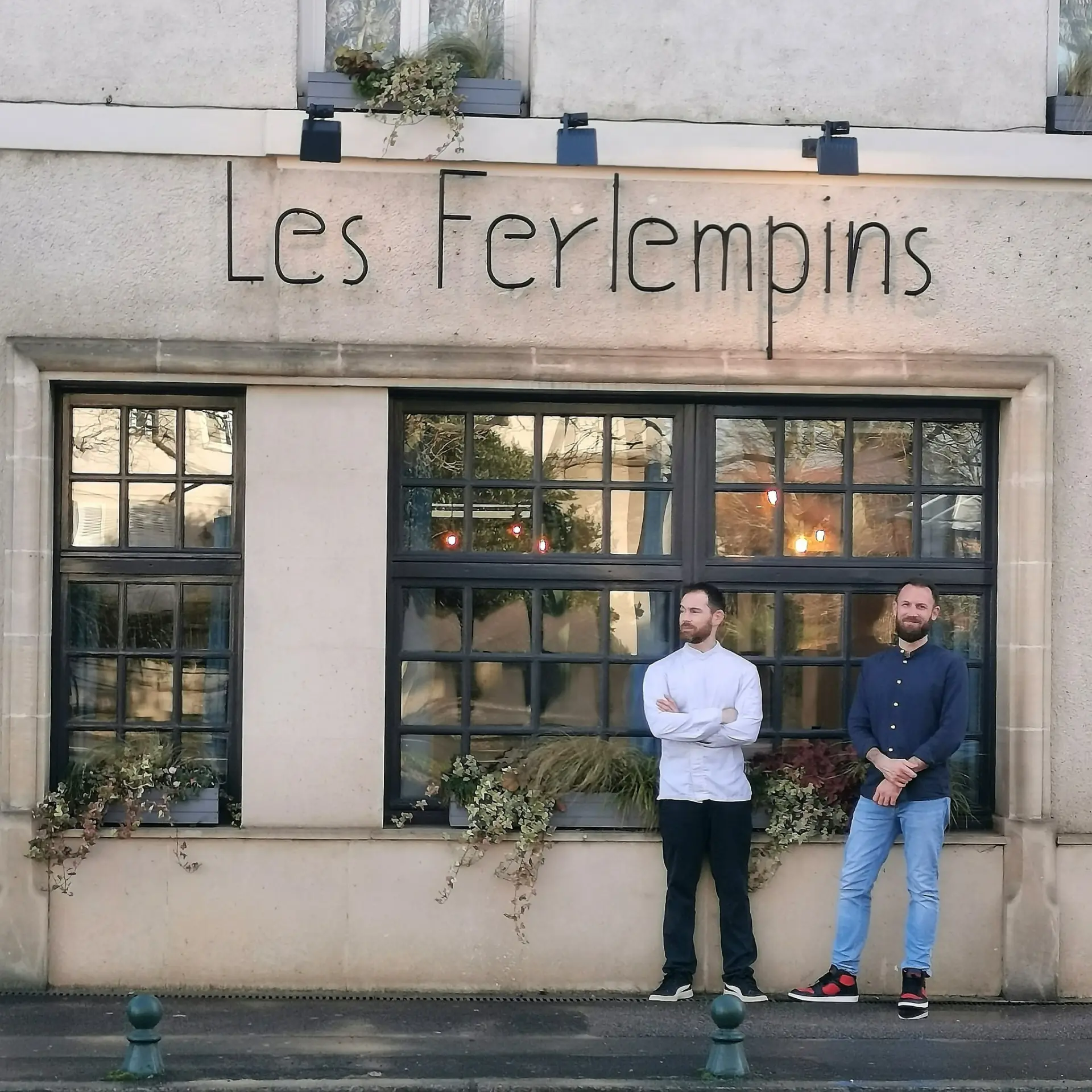 Deux hommes se tiennent devant le restaurant Les Ferlempins, qui possède de grandes fenêtres et des plantes le long de l'extérieur, à quelques pas d'un établissement populaire de Compiègne, comme l'Hôtel de Harlay.