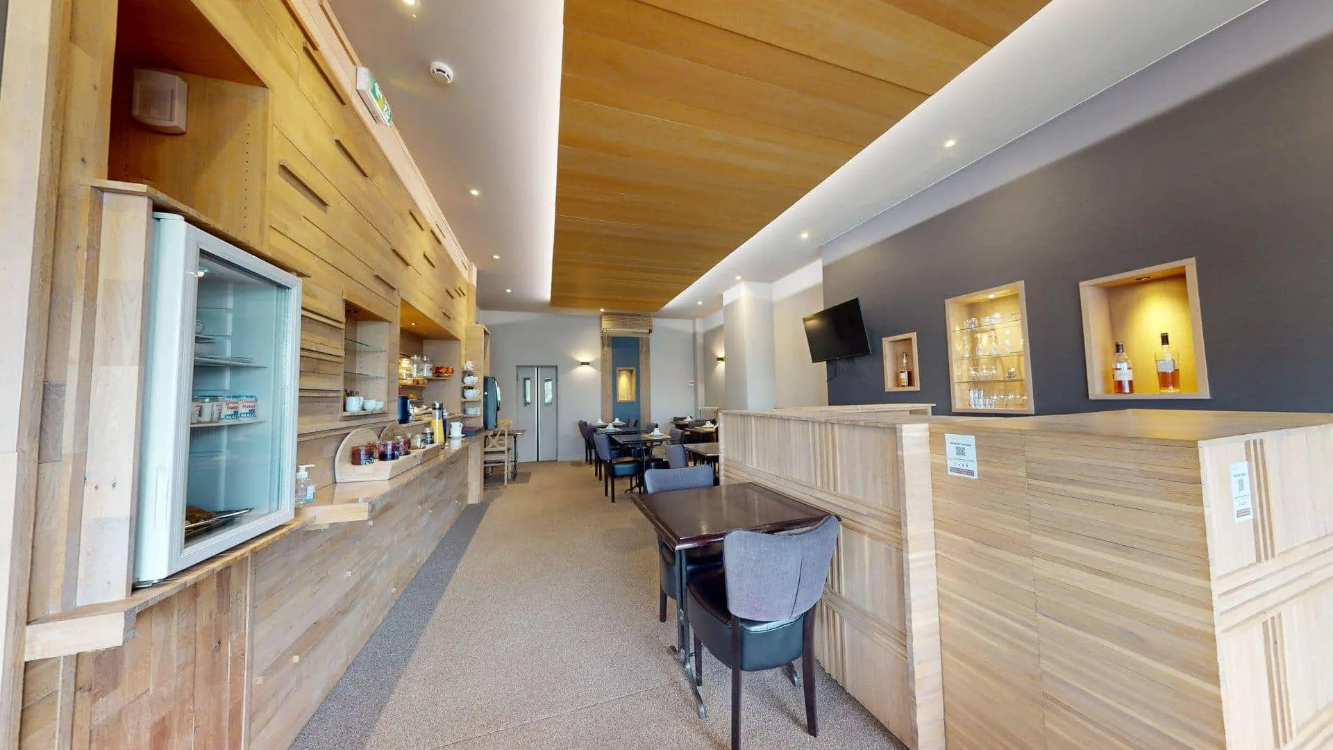 Salon moderne de l'Hôtel de Harlay avec des accents en bois, de petites tables et chaises, une télévision murale, des étagères avec des bouteilles, et un espace snack et boissons en libre-service sur la gauche. Idéal pour se détendre pendant votre séjour à l'hôtel de Compiègne.