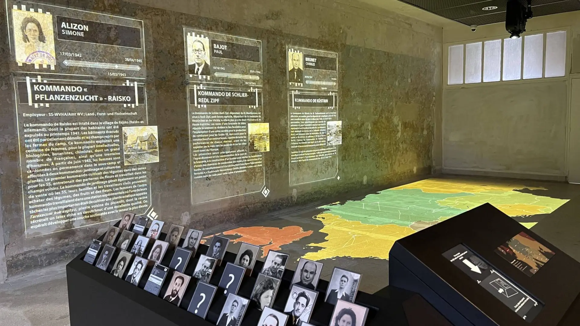 Exposition présentant des biographies projetées, des photographies et des informations sur des personnages historiques sur un mur de l'Hôtel de Harlay, avec au premier plan une carte et un présentoir de cartes de portraits sur une table.