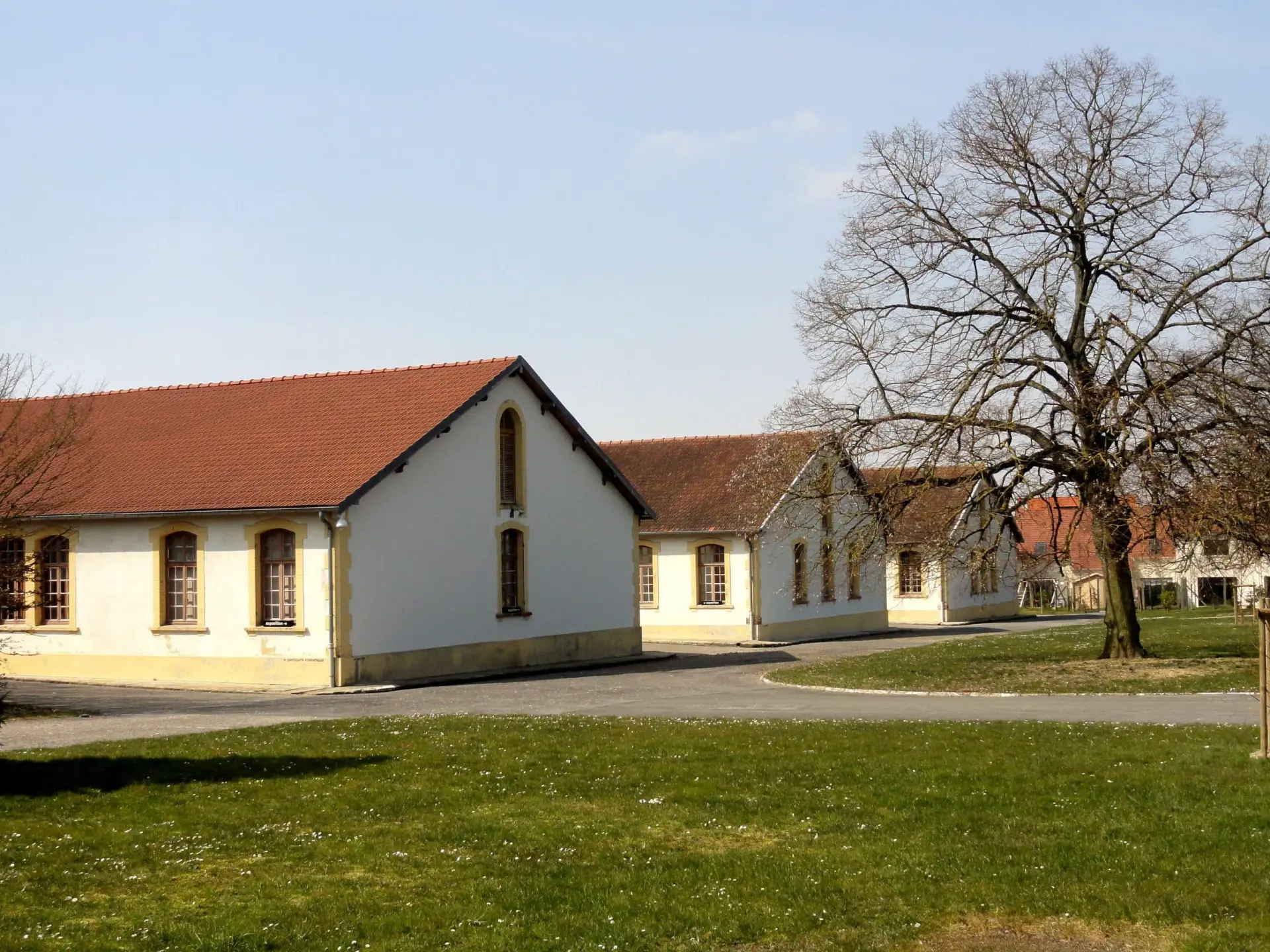 Plusieurs bâtiments blancs de plain-pied aux toits de tuiles rouges sont disposés autour d'une cour pavée de l'Hôtel de Harlay, avec au premier plan un grand arbre sans feuilles et de l'herbe, offrant ainsi le charme de l'Hébergement Compiègne.