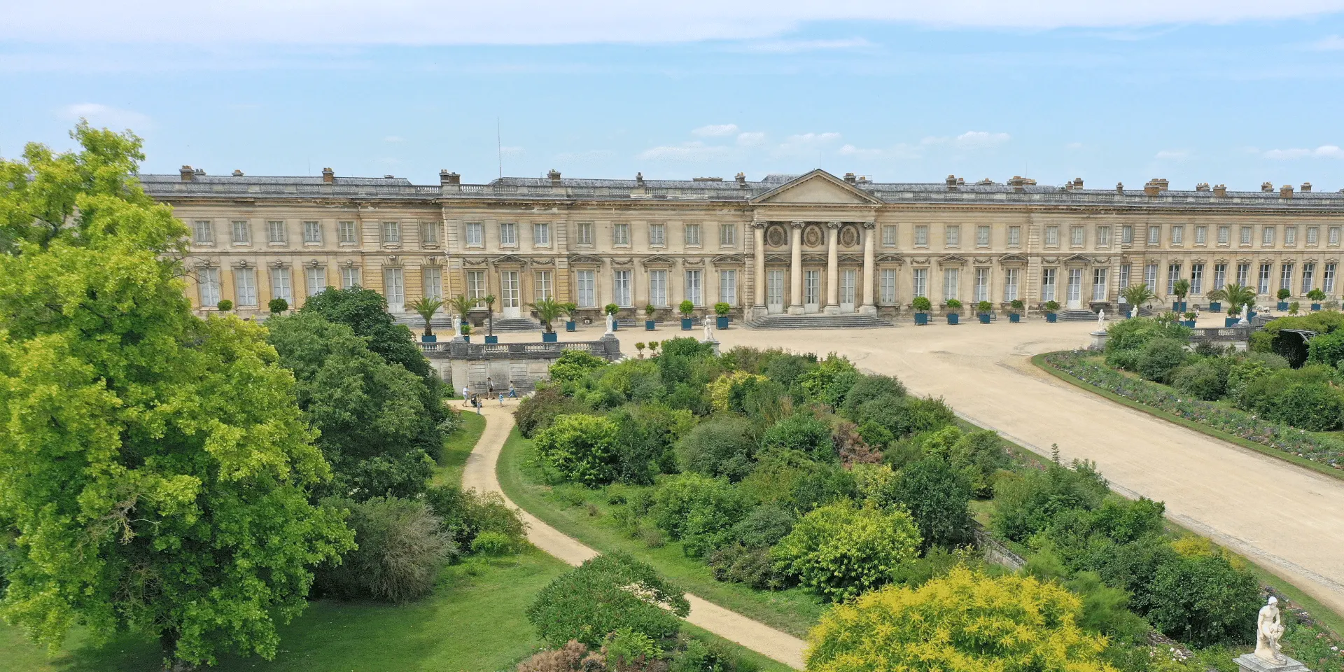 Un grand palais néoclassique avec des colonnes et des fenêtres symétriques se dresse derrière des jardins formels et des allées sous un ciel bleu, offrant une vue pittoresque près de l'Hôtel de Harlay, un hébergement renommé de Compiègne.