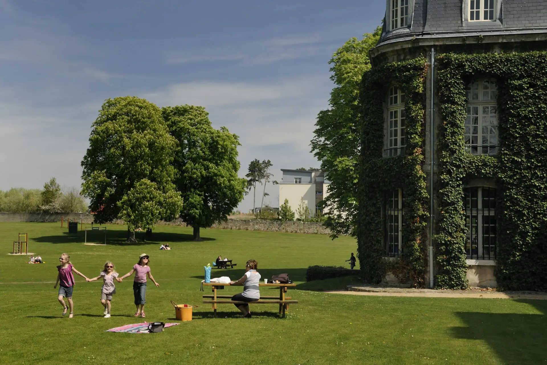 Des enfants se tiennent par la main et marchent sur l'herbe près d'une table de pique-nique, avec un adulte assis à proximité et l'Hôtel de Harlay couvert de lierre à droite. D'autres personnes sont dispersées à l'arrière-plan.