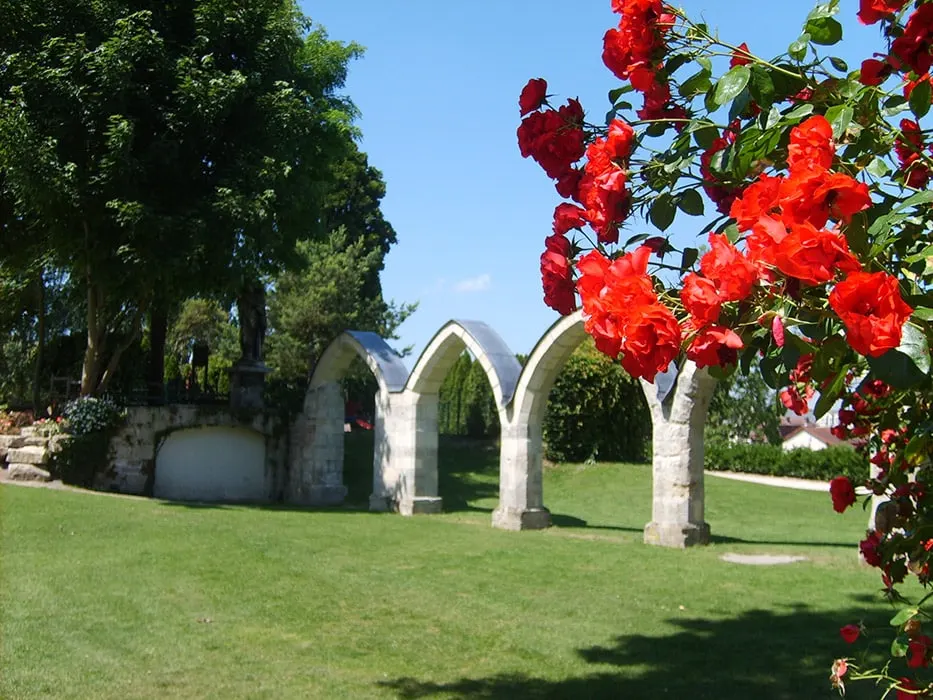 Les ruines d'une arche en pierre se dressent sur une pelouse sous un ciel bleu à l'Hôtel de Harlay, avec des roses d'un rouge éclatant au premier plan à droite.
