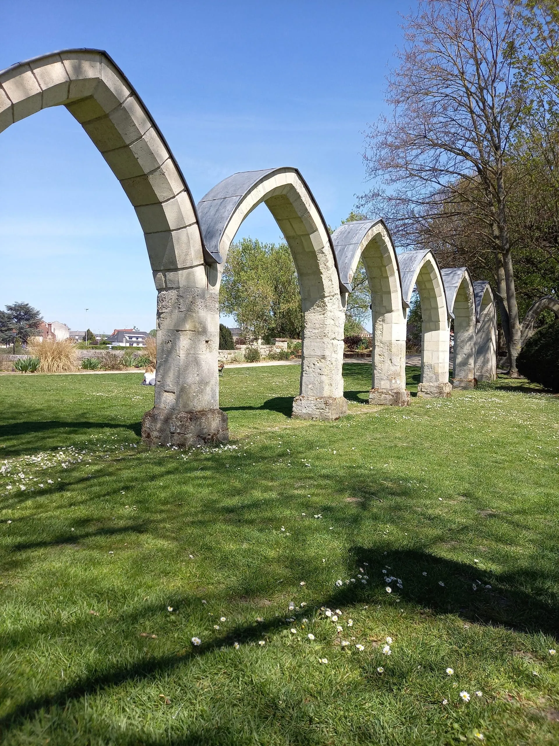 Une rangée de vieilles arches en pierre se dresse sur une pelouse près de l'hôtel historique de Harlay, avec des arbres et des bâtiments en arrière-plan, sous un ciel bleu clair.