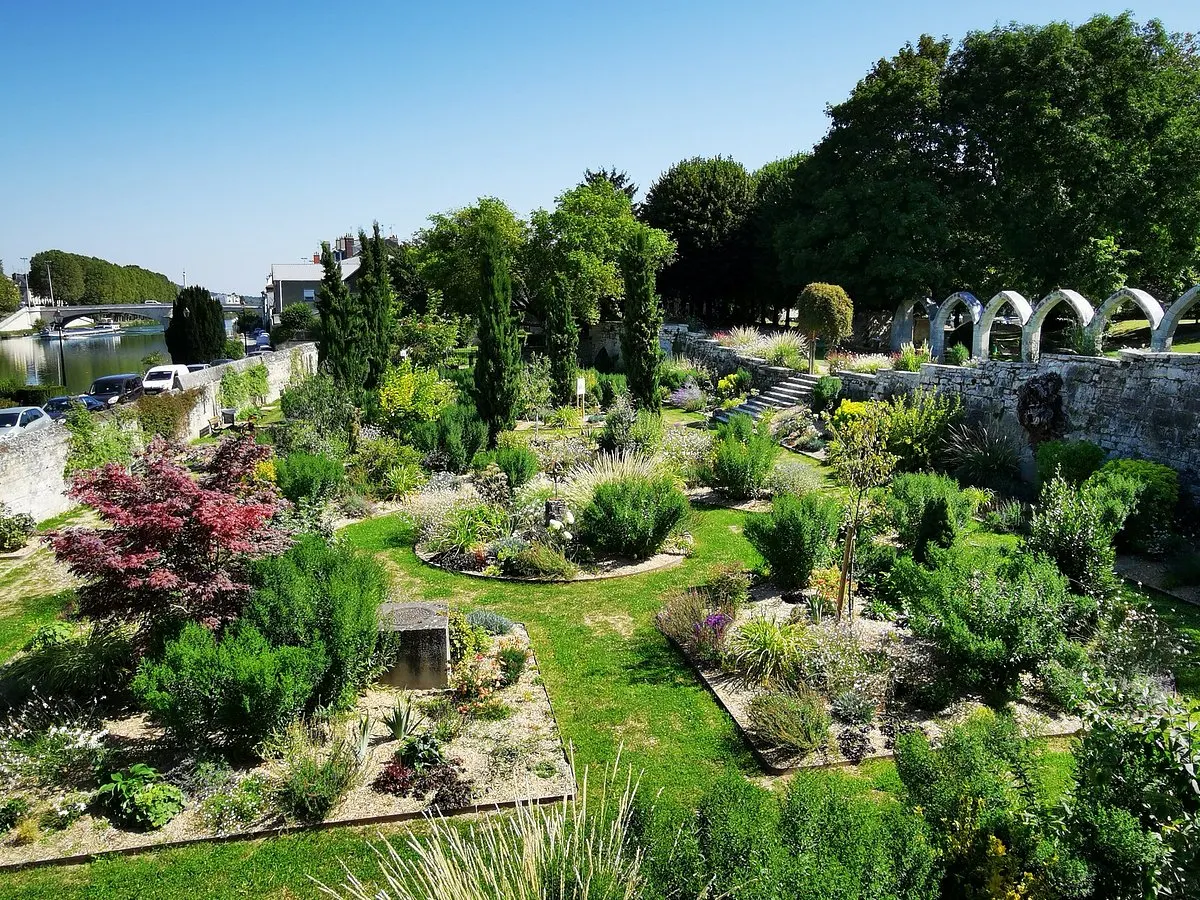 Un jardin botanique bien entretenu avec des parterres de plantes circulaires, des pelouses vertes, une allée en pierre et des arbres offre une échappée paisible près de l'Hôtel de Harlay à Compiègne, encadré par un mur de pierre voûté sous un ciel bleu clair.