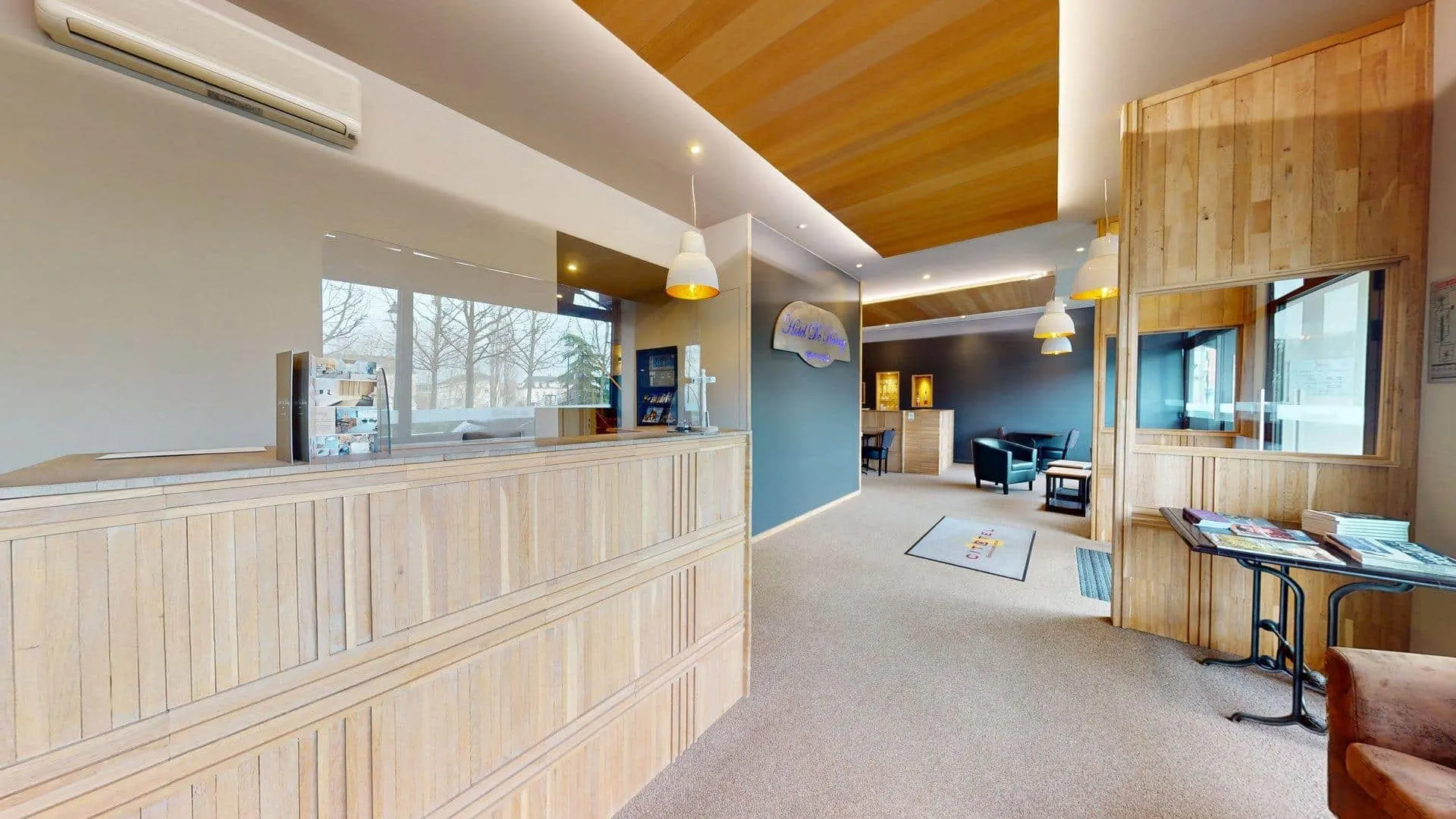 Une réception moderne avec des boiseries, un bureau d'accueil, des sièges en arrière-plan, de grandes fenêtres et des luminaires suspendus, reflétant l'ambiance accueillante de l'Hôtel de Harlay, un hôtel renommé à Compiègne.