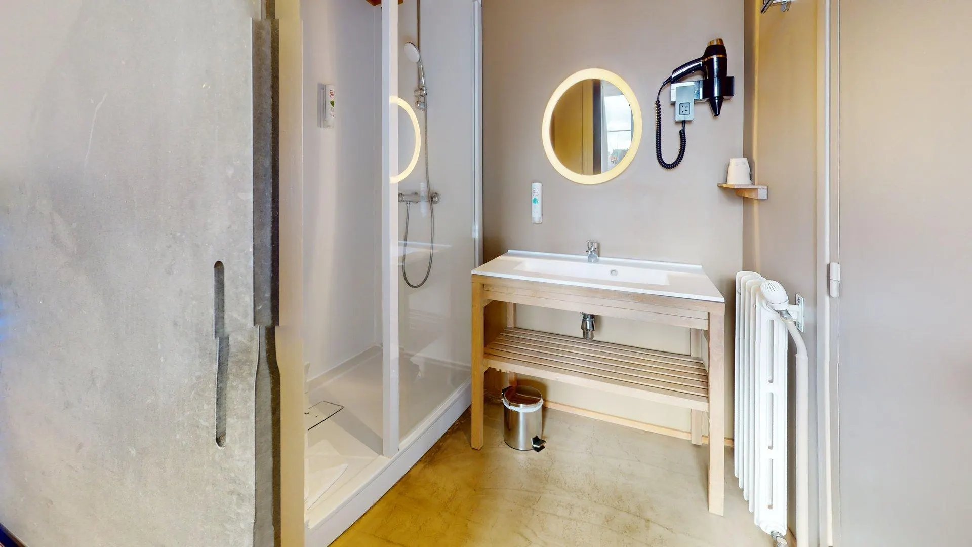 La salle de bains de l'Hôtel de Harlay à Compiègne est équipée d'une douche à l'italienne, d'un meuble en bois avec lavabo, d'un miroir rond, d'un sèche-cheveux mural et d'un radiateur pour votre confort.