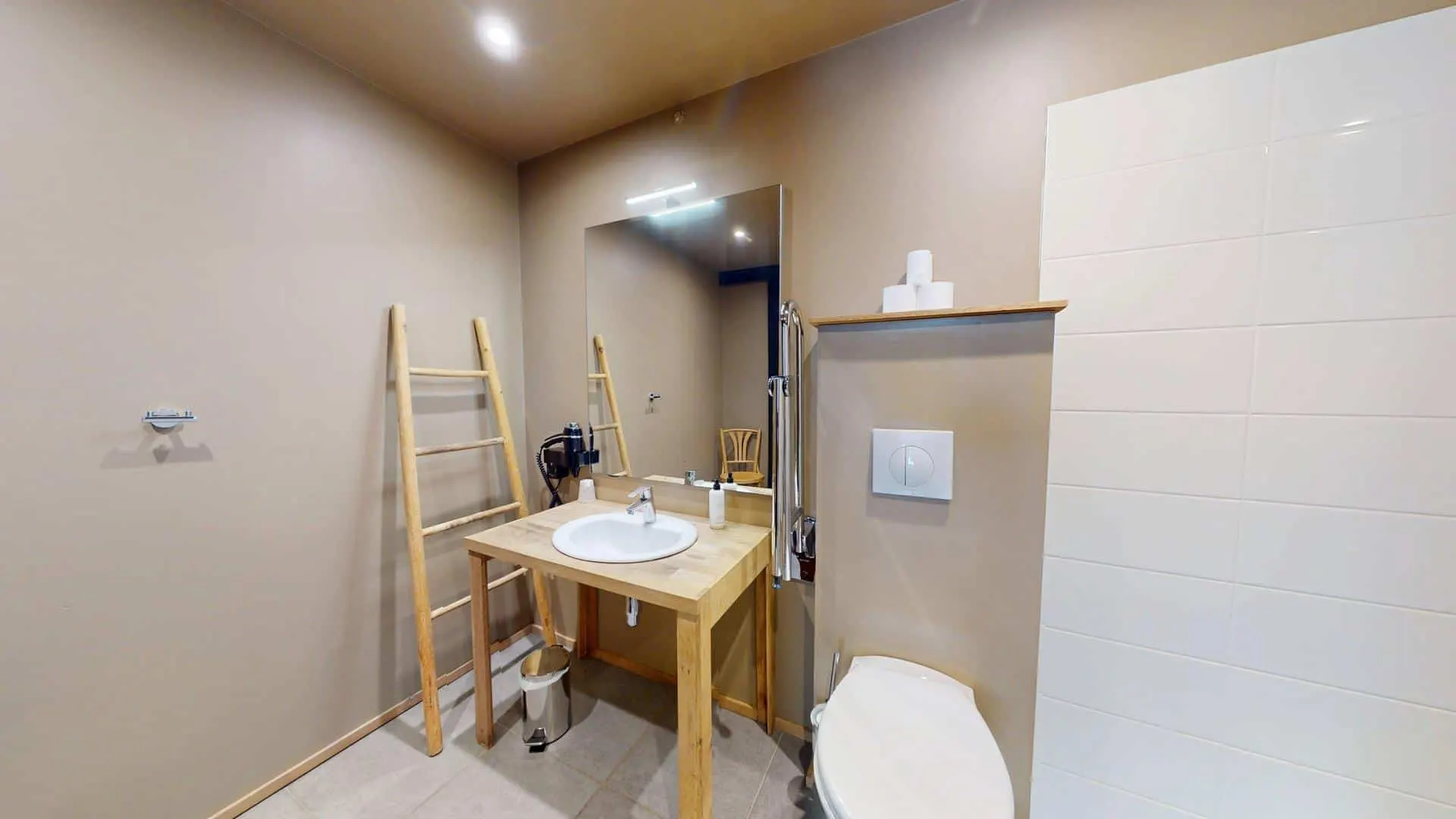 La salle de bains de l'Hôtel de Harlay, avec ses murs beiges, son lavabo en bois, sa vasque ronde, son grand miroir, son porte-serviettes en échelle, ses toilettes et sa petite étagère murale pour le papier hygiénique, offre un confort certain dans cet hébergement de Compiègne.