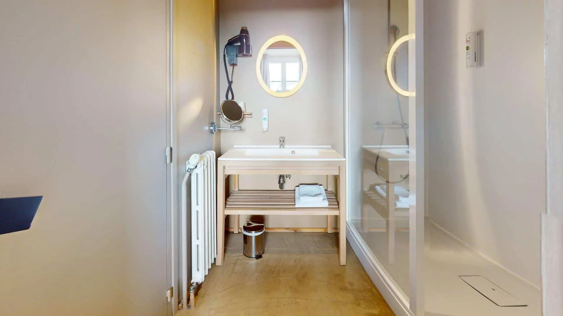 Salle de bain de l'Hôtel de Harlay avec lavabo, miroir, sèche-cheveux mural, serviette sur étagère en bois, poubelle en dessous, et douche avec cloison en verre à droite - idéal pour votre hébergement à Compiègne.