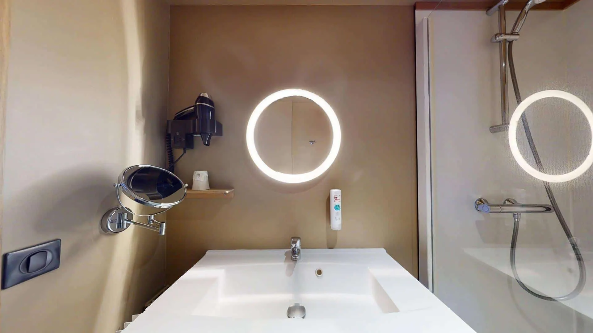 Salle de bain moderne à l'Hôtel de Harlay avec des murs beiges, un lavabo blanc, un miroir rond éclairé, un sèche-cheveux mural et une douche avec une cloison en verre - parfait pour votre séjour à l'Hébergement Compiègne.