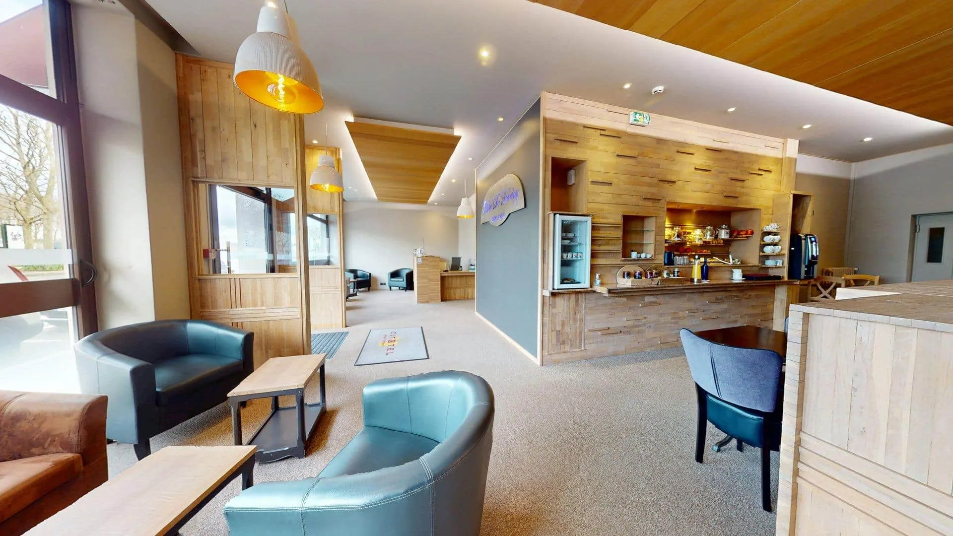 Salon moderne avec des touches de bois, plusieurs fauteuils, de petites tables et un comptoir de bar à café ; de grandes fenêtres offrent une lumière naturelle à l'Hôtel de Harlay, votre hébergement Compiègne idéal.