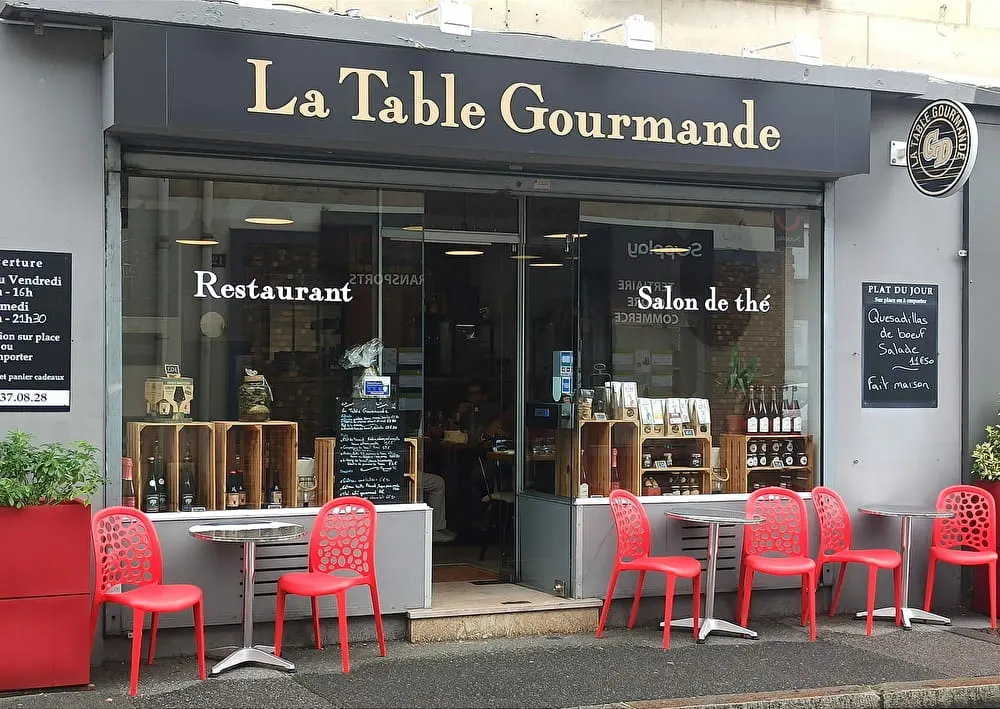 Vue de la rue du restaurant La Table Gourmande avec des chaises et des tables rouges à l'extérieur, une grande vitrine présentant des produits et un menu sur tableau à craie à l'entrée - parfait pour les clients séjournant à l'Hôtel de Harlay ou cherchant Hébergement Compiègne.