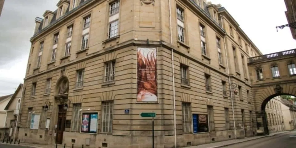 Vue en angle de l'Hôtel de Harlay, un grand bâtiment en pierre beige avec de hautes fenêtres et des bannières sur les murs, situé à l'intersection d'une rue sous un ciel nuageux.