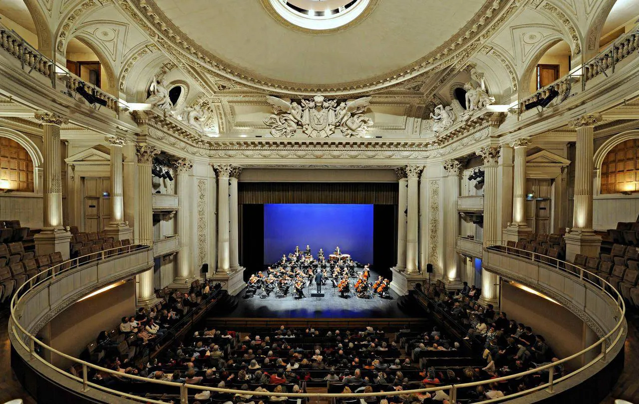 Un orchestre se produit sur scène dans une grande salle de concert ornée d'un plafond en dôme et d'un public assis sur plusieurs niveaux, à quelques pas de l'élégant Hôtel de Harlay, un hébergement de premier choix à Compiègne.