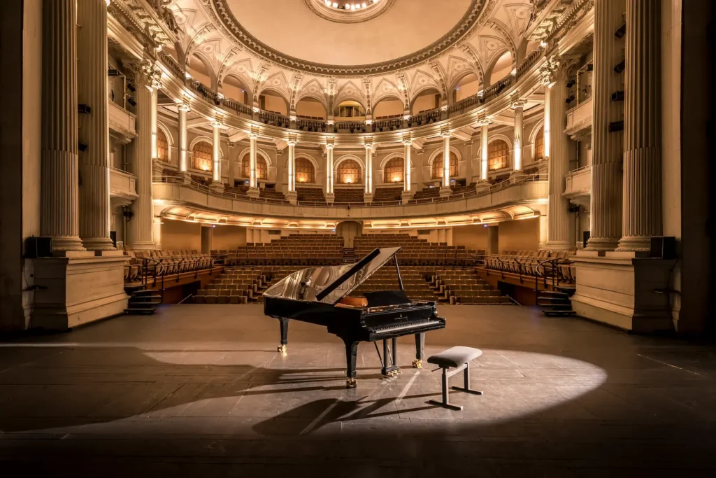 Un piano à queue et son banc sont au centre de la scène, sous les projecteurs, dans une salle de concert vide et ornée, avec des sièges en gradins - une inspiration parfaite pour les clients qui recherchent Hébergement Compiègne près de l'élégant Hôtel de Harlay.
