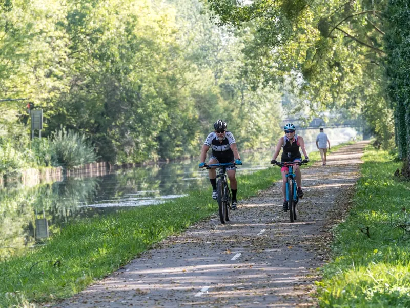 Deux cyclistes roulent sur un chemin bordé d'arbres le long d'un canal près de l'Hôtel de Harlay, avec une personne marchant à l'arrière-plan sous la lumière du soleil.