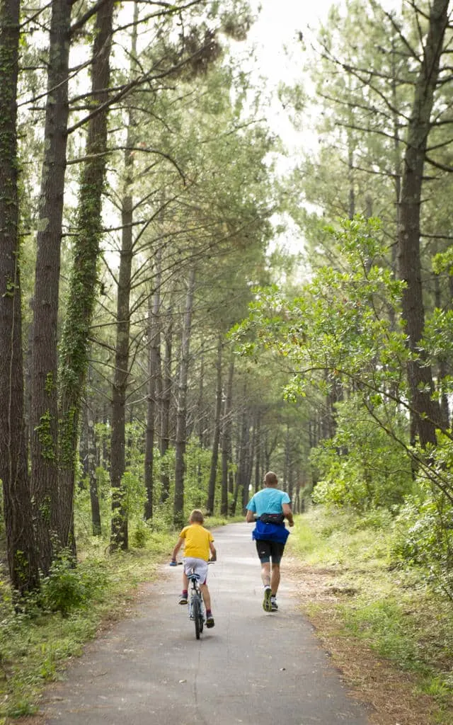 Un adulte fait du jogging et un enfant fait du vélo sur un chemin pavé dans une zone forestière avec de grands arbres, profitant de la nature près d'Hébergement Compiègne.
