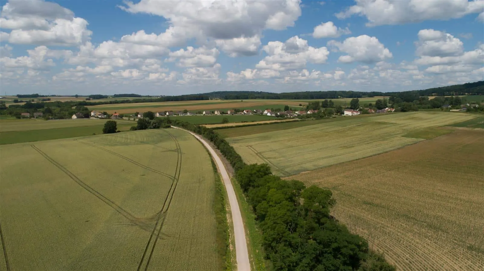 Une route étroite serpente à travers des terres agricoles verdoyantes sous un ciel partiellement nuageux, avec des maisons éparses et des arbres lointains en arrière-plan, évoquant la campagne paisible près de l'hébergement de Compiègne.