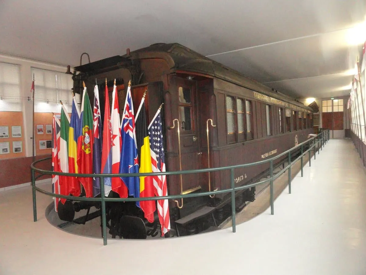 Un wagon historique est exposé à l'intérieur, entouré d'une rambarde et de drapeaux de plusieurs pays, avec des panneaux d'information à proximité - à quelques pas des options d'Hébergement Compiègne comme l'Hôtel de Harlay pour un séjour mémorable.